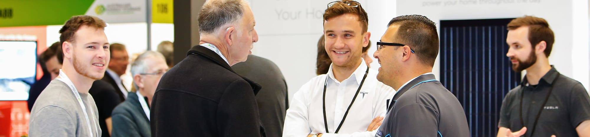 Tesla staff chatting with visitors in front of a Powerwall display at the Australian Energy Storage exhibition, banner crop